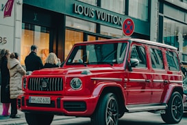 A bright red luxury SUV is parked on a city street in front of a Louis Vuitton store. Several people are standing nearby, dressed in winter clothing, suggesting cooler weather. The scene has a high-end, urban atmosphere with a mix of pedestrians and high-end stores.