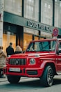 A luxury red SUV is parked on a city street, in front of a Louis Vuitton store. Several people are standing nearby, some appearing to look into the store windows. The storefront features large glass windows and a prominent brand logo. The weather appears clear, suggesting a daytime setting.