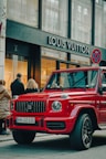 A luxury red SUV is parked on a city street, in front of a Louis Vuitton store. Several people are standing nearby, some appearing to look into the store windows. The storefront features large glass windows and a prominent brand logo. The weather appears clear, suggesting a daytime setting.
