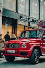 A luxury red SUV is parked on a city street, in front of a Louis Vuitton store. Several people are standing nearby, some appearing to look into the store windows. The storefront features large glass windows and a prominent brand logo. The weather appears clear, suggesting a daytime setting.