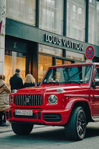 A luxury red SUV is parked on a city street, in front of a Louis Vuitton store. Several people are standing nearby, some appearing to look into the store windows. The storefront features large glass windows and a prominent brand logo. The weather appears clear, suggesting a daytime setting.