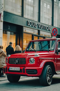 A luxury red SUV is parked on a city street, in front of a Louis Vuitton store. Several people are standing nearby, some appearing to look into the store windows. The storefront features large glass windows and a prominent brand logo. The weather appears clear, suggesting a daytime setting.