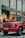 A luxury red SUV is parked on a city street, in front of a Louis Vuitton store. Several people are standing nearby, some appearing to look into the store windows. The storefront features large glass windows and a prominent brand logo. The weather appears clear, suggesting a daytime setting.