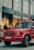 A luxury red SUV is parked on a city street, in front of a Louis Vuitton store. Several people are standing nearby, some appearing to look into the store windows. The storefront features large glass windows and a prominent brand logo. The weather appears clear, suggesting a daytime setting.