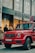 A luxury red SUV is parked on a city street, in front of a Louis Vuitton store. Several people are standing nearby, some appearing to look into the store windows. The storefront features large glass windows and a prominent brand logo. The weather appears clear, suggesting a daytime setting.