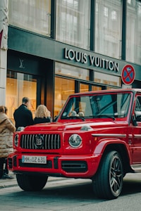 A luxury red SUV is parked on a city street, in front of a Louis Vuitton store. Several people are standing nearby, some appearing to look into the store windows. The storefront features large glass windows and a prominent brand logo. The weather appears clear, suggesting a daytime setting.