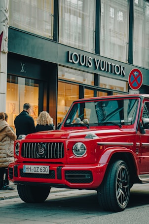 A luxury red SUV is parked on a city street, in front of a Louis Vuitton store. Several people are standing nearby, some appearing to look into the store windows. The storefront features large glass windows and a prominent brand logo. The weather appears clear, suggesting a daytime setting.