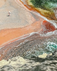 A person wearing a hat and beachwear walks along a colorful beach with striking red and orange sand meeting the blue-green ocean waves. The shoreline creates an interesting pattern as the water blends with the vibrant sands, surrounded by natural rocky formations.
