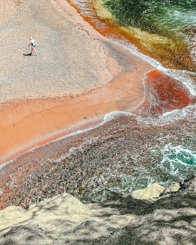 A person wearing a hat and beachwear walks along a colorful beach with striking red and orange sand meeting the blue-green ocean waves. The shoreline creates an interesting pattern as the water blends with the vibrant sands, surrounded by natural rocky formations.