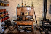 An old-fashioned kitchen with a wooden hutch filled with various jars and kitchen utensils. Shelves on the left are lined with colorful jars of preserved foods. A vintage stove stands on the right, surrounded by pots and containers. The wooden floor and rustic walls add to the antique ambiance.