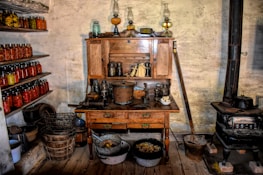 An old-fashioned kitchen with a wooden hutch filled with various jars and kitchen utensils. Shelves on the left are lined with colorful jars of preserved foods. A vintage stove stands on the right, surrounded by pots and containers. The wooden floor and rustic walls add to the antique ambiance.