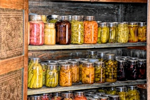 Multiple shelves filled with numerous jars of preserved vegetables and pickles inside what appears to be a wooden pantry or storage area. The jars contain a variety of colors and types of vegetables.