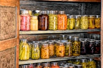 Multiple shelves filled with numerous jars of preserved vegetables and pickles inside what appears to be a wooden pantry or storage area. The jars contain a variety of colors and types of vegetables.