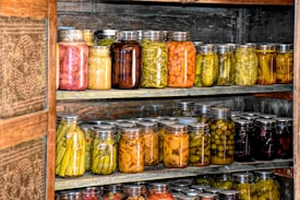 Multiple shelves filled with numerous jars of preserved vegetables and pickles inside what appears to be a wooden pantry or storage area. The jars contain a variety of colors and types of vegetables.