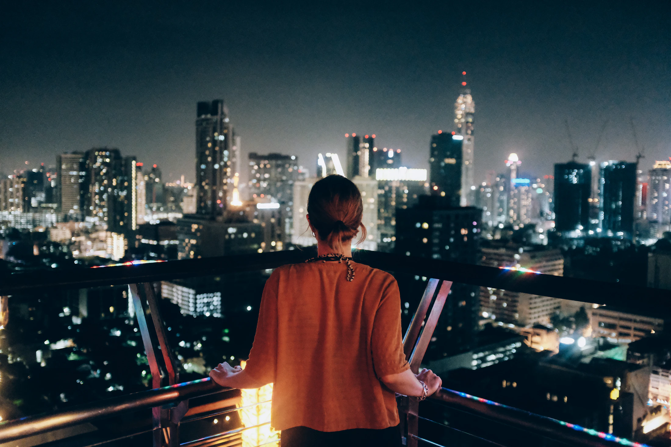 An anonymous figure standing on a high-rise balcony at dawn, resting on the railing while looking out over a city skyline stretching into the distance