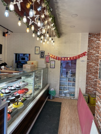 A bakery interior is adorned with festive decorations, including hanging lights and star-shaped ornaments. A glass display case is filled with various cakes, featuring colorful frosting and decorations. A 'Merry Christmas' banner is displayed on the wall. Framed pictures and a menu are mounted above the display. The space has a warm and cozy ambiance with white brick walls and wooden flooring.