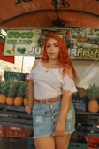 A woman with long red hair is standing in front of a market stall. She is wearing a white off-shoulder top, a denim skirt, and a brown belt. Behind her are stacks of pineapples and a sign with the text 'COCO GELADO JUICER FRUTAS'. The market setting is informal with colorful fruit on display.