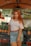 A woman with long red hair is standing in front of a market stall. She is wearing a white off-shoulder top, a denim skirt, and a brown belt. Behind her are stacks of pineapples and a sign with the text 'COCO GELADO JUICER FRUTAS'. The market setting is informal with colorful fruit on display.