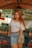A woman with long red hair is standing in front of a market stall. She is wearing a white off-shoulder top, a denim skirt, and a brown belt. Behind her are stacks of pineapples and a sign with the text 'COCO GELADO JUICER FRUTAS'. The market setting is informal with colorful fruit on display.