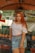A woman with long red hair is standing in front of a market stall. She is wearing a white off-shoulder top, a denim skirt, and a brown belt. Behind her are stacks of pineapples and a sign with the text 'COCO GELADO JUICER FRUTAS'. The market setting is informal with colorful fruit on display.