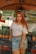 A woman with long red hair is standing in front of a market stall. She is wearing a white off-shoulder top, a denim skirt, and a brown belt. Behind her are stacks of pineapples and a sign with the text 'COCO GELADO JUICER FRUTAS'. The market setting is informal with colorful fruit on display.