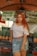 A woman with long red hair is standing in front of a market stall. She is wearing a white off-shoulder top, a denim skirt, and a brown belt. Behind her are stacks of pineapples and a sign with the text 'COCO GELADO JUICER FRUTAS'. The market setting is informal with colorful fruit on display.