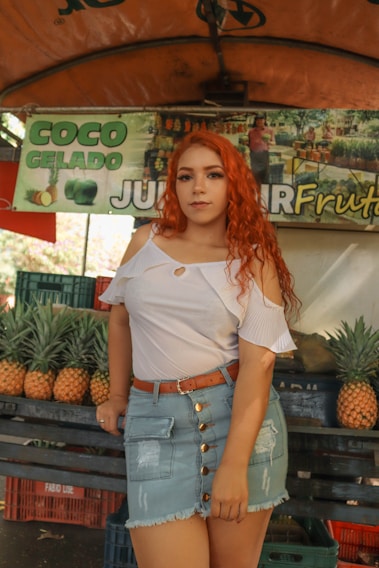A woman with long red hair is standing in front of a market stall. She is wearing a white off-shoulder top, a denim skirt, and a brown belt. Behind her are stacks of pineapples and a sign with the text 'COCO GELADO JUICER FRUTAS'. The market setting is informal with colorful fruit on display.