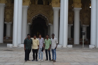 Five young men stand together in front of a building with tall, ornate columns and intricate architectural details. The ground is paved with stone tiles, and the backdrop features dark, arched openings.