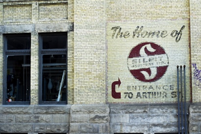A brick wall with a large sign painted on it displaying the name 'Silco Industries Ltd.' along with an arrow pointing towards the entrance at 70 Arthur St. The wall is part of an older building with two large windows and visible pipes, adding an industrial vibe.