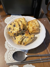 A white plate contains several freshly baked scones with visible chocolate chips, placed on a wooden table adorned with a white lace doily. In front of the plate, there is a metal spoon and a set of chopsticks. A black appliance is partially visible in the background.