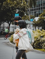 Two people, wearing masks, walk side by side on a city street, with one person holding a small dog wrapped in a fluffy jacket. They carry a turquoise pet backpack with a transparent dome. In the background, there's a cyclist wearing green, trees, and a modern building with glass windows. A sign indicating a sale is visible, hanging on a lamppost.