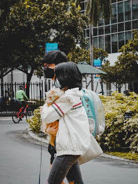 Two people, wearing masks, walk side by side on a city street, with one person holding a small dog wrapped in a fluffy jacket. They carry a turquoise pet backpack with a transparent dome. In the background, there's a cyclist wearing green, trees, and a modern building with glass windows. A sign indicating a sale is visible, hanging on a lamppost.
