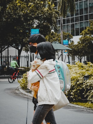 Two people, wearing masks, walk side by side on a city street, with one person holding a small dog wrapped in a fluffy jacket. They carry a turquoise pet backpack with a transparent dome. In the background, there's a cyclist wearing green, trees, and a modern building with glass windows. A sign indicating a sale is visible, hanging on a lamppost.