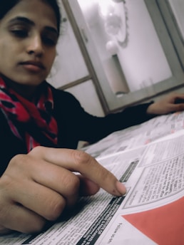 A person sitting at a table, focusing on reading a newspaper, with their finger pointing at specific text. The individual is wearing a red and black scarf, and the room appears to have a soft, neutral decor.