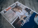 A newspaper titled 'Delhi Times' is placed on a wooden surface. The front page features an advertisement with a woman in a white blouse, leaning her arm on a surface. A watch advertisement is prominently displayed with the brand 'Longines'. The ad includes the text 'Elegance is an attitude' accompanied by a signature.