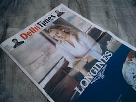 A newspaper titled 'Delhi Times' is placed on a wooden surface. The front page features an advertisement with a woman in a white blouse, leaning her arm on a surface. A watch advertisement is prominently displayed with the brand 'Longines'. The ad includes the text 'Elegance is an attitude' accompanied by a signature.