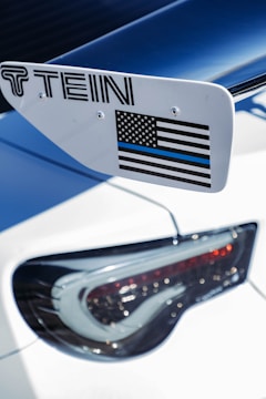 A close-up view of a car's rear section, featuring a spoiler with a 'TEIN' logo and an American flag decal in black and white with a blue line. The tail light is sleek and modern, characterized by intricate detailing.