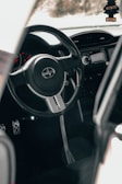 The interior view of a car focusing on the steering wheel with a Scion logo. The dashboard and a part of the car’s odometer are visible, along with metallic pedals. A rearview mirror accessory is hanging, shaped like a tree with text on it.