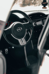 The interior view of a car focusing on the steering wheel with a Scion logo. The dashboard and a part of the car’s odometer are visible, along with metallic pedals. A rearview mirror accessory is hanging, shaped like a tree with text on it.