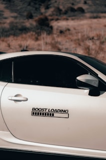 A sleek silver car is parked against a backdrop of dry, grassy hills. The car's door features a sticker that reads 'Boost Loading' with a progress bar below it, adding a playful touch.