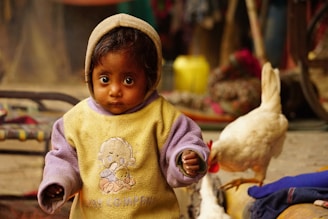 A young child with large, expressive eyes is wearing a warm, hooded sweater featuring a cartoon character. The child stands in an indoor setting, with a chicken nearby. The background is slightly blurred, showing household items and colorful textiles.