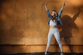 A woman wearing a denim jacket and white athletic wear is standing with her arms raised against a textured orange wall. Her shadow is visible on the wall, creating an artistic effect.
