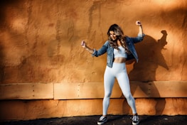 A woman in athletic wear poses energetically against a textured orange wall, casting a shadow. She is wearing white leggings, a white crop top, a denim jacket, and black sneakers.