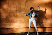 A woman in athletic wear poses energetically against a textured orange wall, casting a shadow. She is wearing white leggings, a white crop top, a denim jacket, and black sneakers.