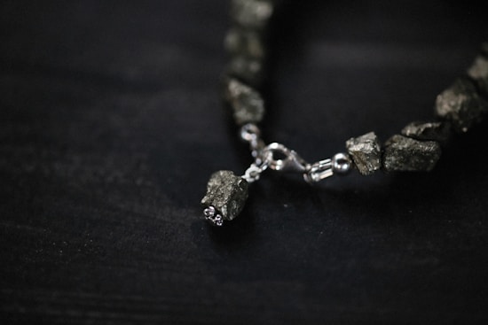 A close-up view of a bracelet featuring irregularly shaped metallic stones and a shiny metal clasp. The stones have a rough texture and give an earthy yet refined appearance. The background is a dark surface, emphasizing the subtle shine of the stones and clasp.