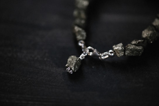 A close-up view of a bracelet featuring irregularly shaped metallic stones and a shiny metal clasp. The stones have a rough texture and give an earthy yet refined appearance. The background is a dark surface, emphasizing the subtle shine of the stones and clasp.