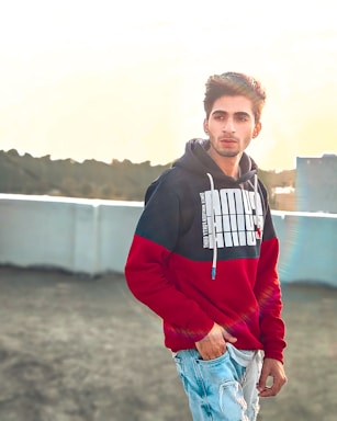 A young person stands outdoors wearing a red and black hoodie with graphic text on it, paired with light blue ripped jeans. The individual has a casual expression and is positioned against a bright sky background with sunlight casting a subtle flare.