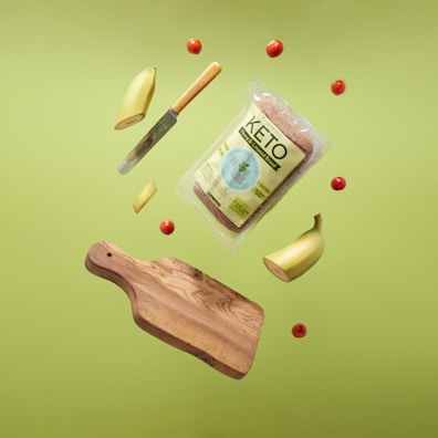 A levitating display featuring a wooden cutting board, a sliced banana, small red tomatoes, a bread package labeled 'Keto', and a knife with a wooden handle, all set against a bright green background.