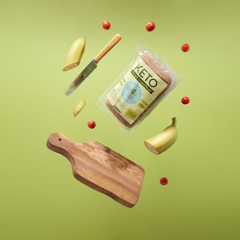 A levitating display featuring a wooden cutting board, a sliced banana, small red tomatoes, a bread package labeled 'Keto', and a knife with a wooden handle, all set against a bright green background.