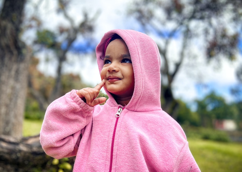 a little girl in a pink jacket standing in front of a tree